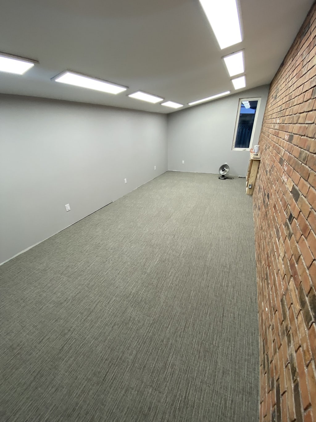Finished lower-level room with grey walls, carpet, and an exposed brick wall